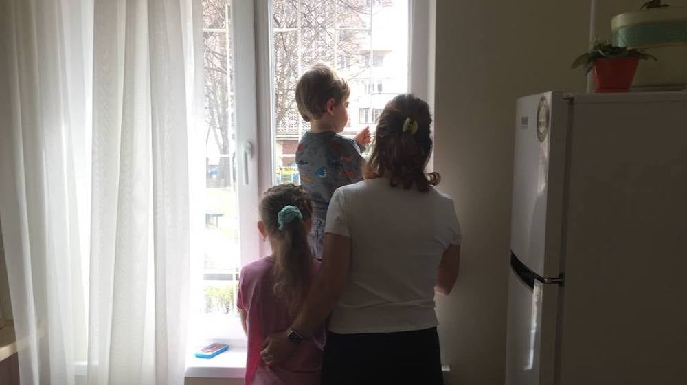 A woman stands in the shadows with two children, looking out a window