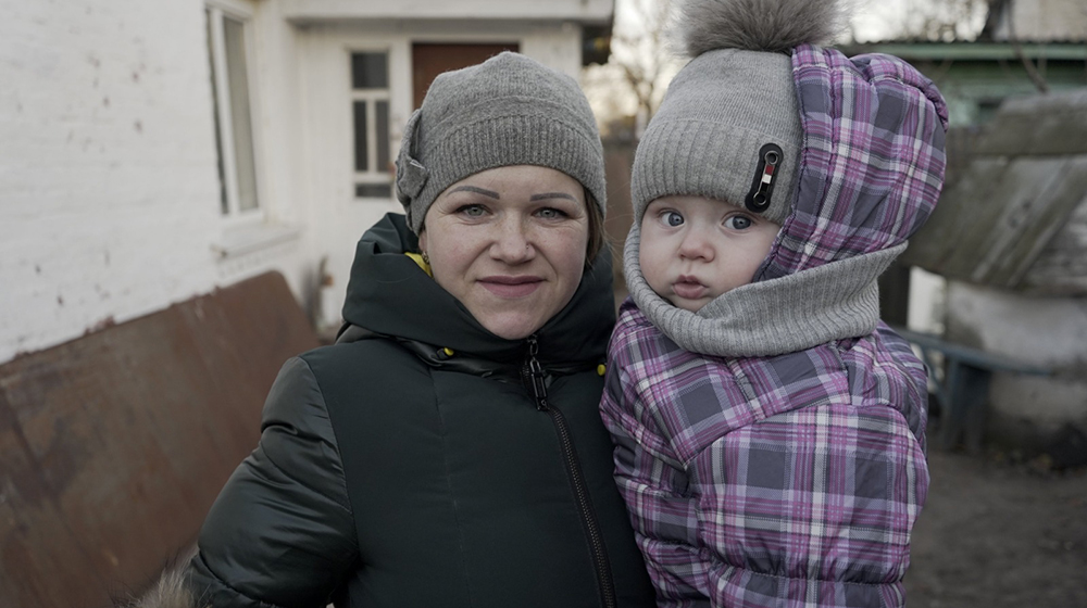 A mother and her child stand outdoors. They are wearing jackets and winter hats, and looking into the camera. 