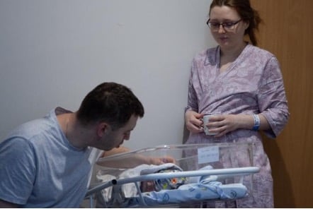 New mother Oleksandra Chebotar stands and looks down at her husband Oleksandr touching their newborn baby in the hospital crib
