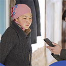 A woman wearing a pink scarf around her hair looks at a cell phone being held by another person