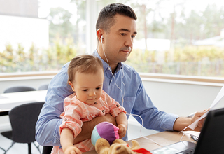 A man in a blue shirt sits at a desk. He is holding a baby girl and looking at a stack of papers in his hand.