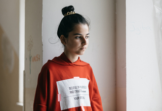 A young girl is standing a room with white walls. She is wearing a red hoodie with a slogan on, her hair is tied in a bun.
