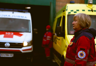 A woman with short blonde hair wearing a red medical jacket stands in front of ambulances
