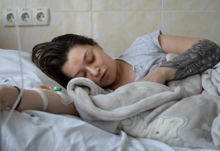 A woman lays in a hospital bed with her eyes closed. Next to her is a blanket wrapped up around a baby.