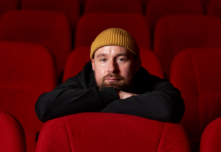 Bohdan Zhuk in a yellow beanie sits in a red movie theatre seat, leaning forward with his arms crossed over the top of the seat