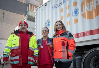 A man and two women, wearing medical clothes, stand outside in front of a truck. They are looking into the camera.