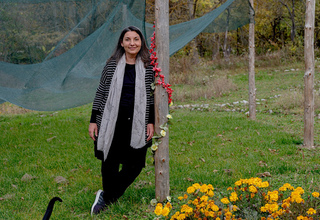 A woman stands in a grassy yard, leaning against a pole and smiling into the camera