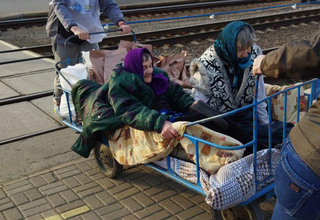 Two older women are sitting on a cart at a train station in Ukraine. A man is pushing the cart from behind.