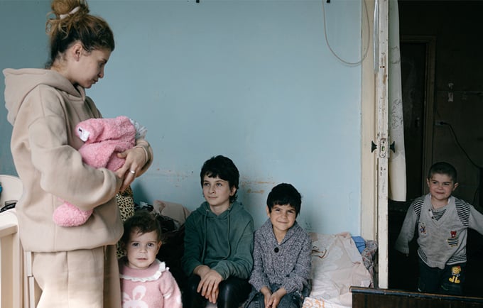A woman is standing holding a baby. Four young children are also in the room. They are all smiling.