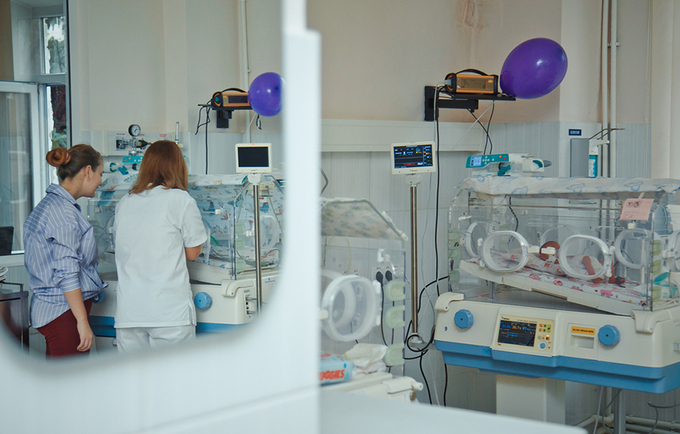 A maternity ward seen through an opened door. Three incubators are visible and a nurse & a woman with their backs to the camera.