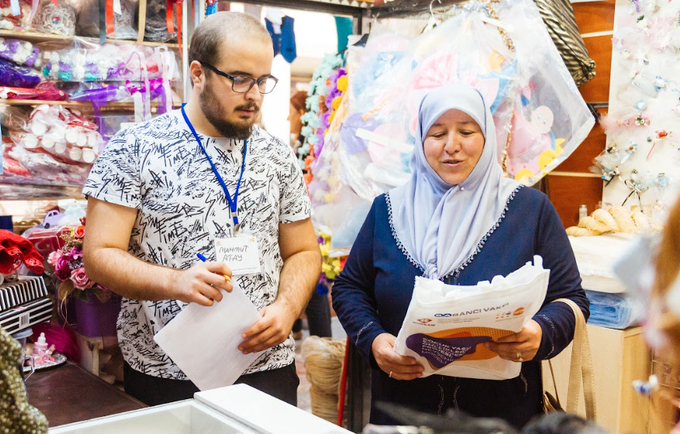 A woman is talking to a clothes seller in his shop. He is wearing glasses. She is wearing a head covering.