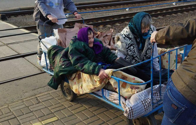 Two older women are sitting on a cart at a train station in Ukraine. A man is pushing the cart from behind.