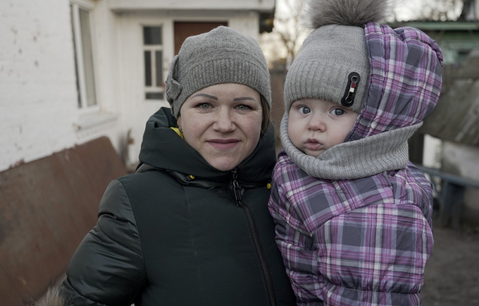 A mother and her child stand outdoors. They are wearing jackets and winter hats, and looking into the camera. 