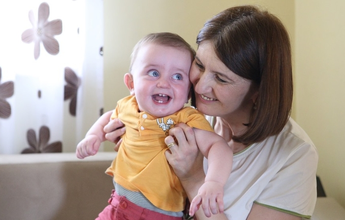 A woman with brown hair is holding a baby. The baby is wearing a yellow top. They are both smiling. 