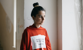 A young girl is standing a room with white walls. She is wearing a red hoodie with a slogan on, her hair is tied in a bun.
