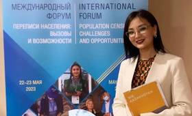 A woman is standing in front of a banner. She is wearing glasses and smiling and she is holding a folder.