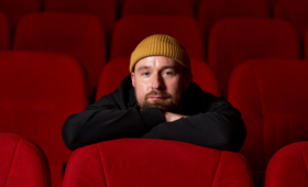 Bohdan Zhuk in a yellow beanie sits in a red movie theatre seat, leaning forward with his arms crossed over the top of the seat