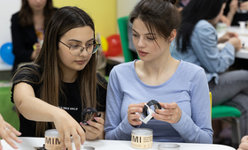 Two young women sit at a desk in a classroom, looking at watches