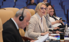Woman with blonde hair and glasses speaks at conference while man with headphones listens in foreground