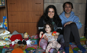 A woman, man, and girl child sit on the floor of a house. All three are smiling and are surrounded by toys and household items.