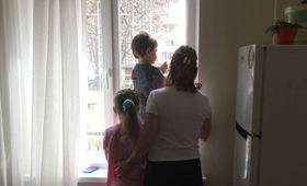 A woman stands in the shadows with two children, looking out a window