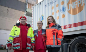 A man and two women, wearing medical clothes, stand outside in front of a truck. They are looking into the camera.