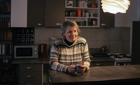 An older woman is sitting at a table in a kitchen. She has her hands around a drinking cup. She is smiling.