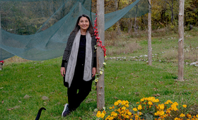 A woman stands in a grassy yard, leaning against a pole and smiling into the camera