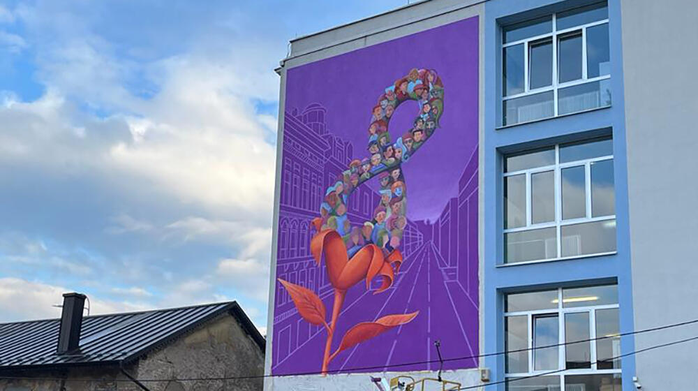 A blue and white building is shown with a large purple mural painted on the side. The mural has a flower with an 8 inside of it.