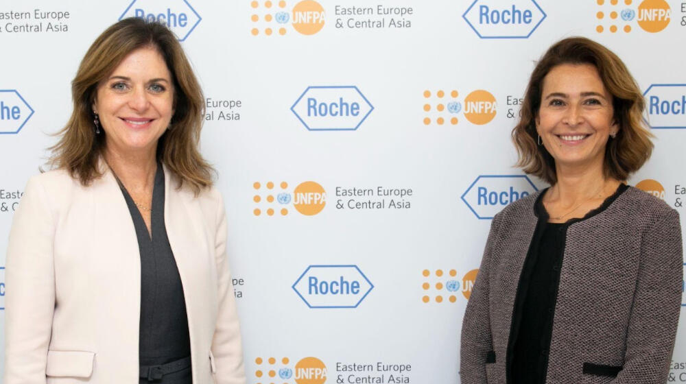 Two women stand smiling in front of a white screen with logos on it