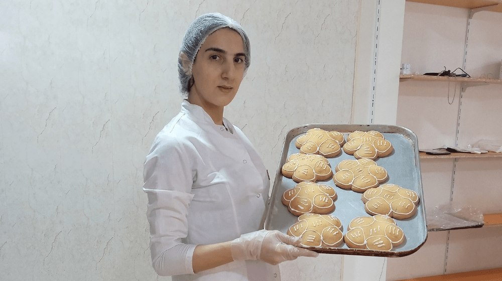 Young woman wearing white chef's jacket, hair net and plastic gloves holds up sheet pan filled with flower-shaped backed goods with white decorative icing on top