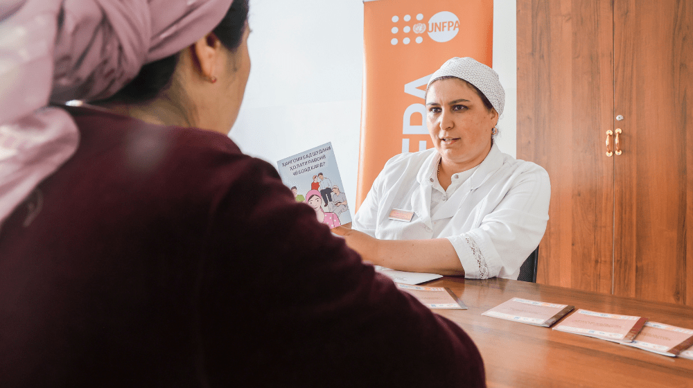 Two women sit at a wooden table inside a room. The camera looks over the right shoulder of one woman wearing a dark maroon sweater and a light purple headscarf. Across from her a woman in white doctor's uniform and cap speaks to her while holding an information brochure. An orange UNFPA banner is in the background