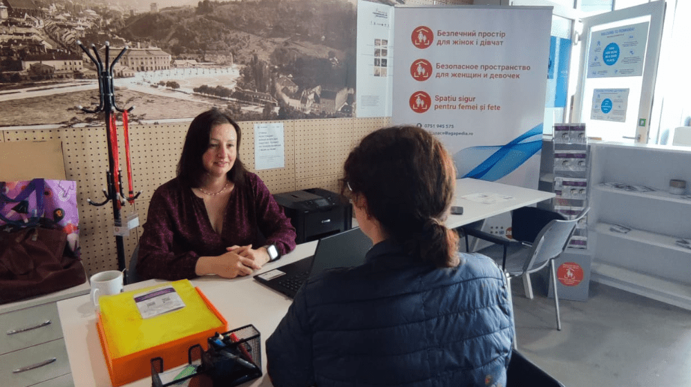 Two women sit at a desk facing each other inside the Safe Space office lobby, one woman with dark brown hair in a low ponytail and black jackets has her back to the camera. The woman across from her greets her with a welcoming smile. A UNFPA Safe Space banner stands in the background.