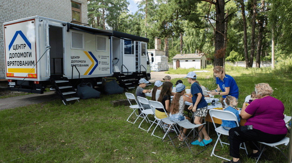 Two adults supervise a group of children playing at a table set up outside on the grass in front of a mobile support centre truck