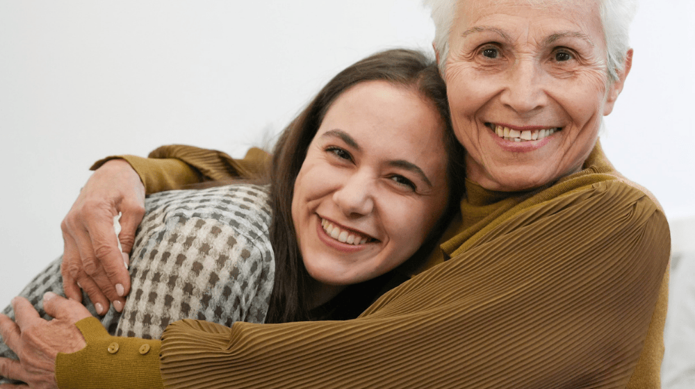 Close up of an older woman with short white hair and a caramel-coloured blouse smiling and wrapping her arms around a younger woman who is also smiling widely at the camera, leaning into the older woman's hug. The young woman has long brown hair and is wearing a green-and-white checked top