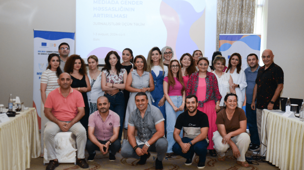 Large group photo of dozens of journalists, bloggers and editors smiling at the camera, mostly standing but some sitting and kneeling in the front row, between long meeting room tables and in front of a projector screen showing a media training presentation. 