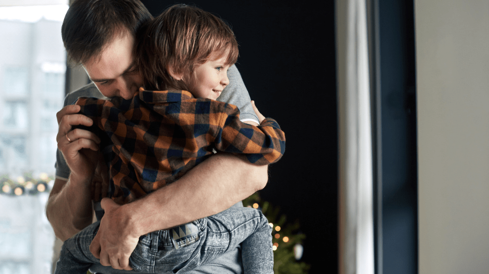 A father in a grey tshirt and brown hair carries his toddler son in his arms and hugs him. The son is wearing an orange and blue plaid collared shirt and has brown shaggy hair. The father's face is nuzzled close to the boy's neck. The boy's back is to the camera, but his face can be seen smiling in side profile. The background appears to be their home.