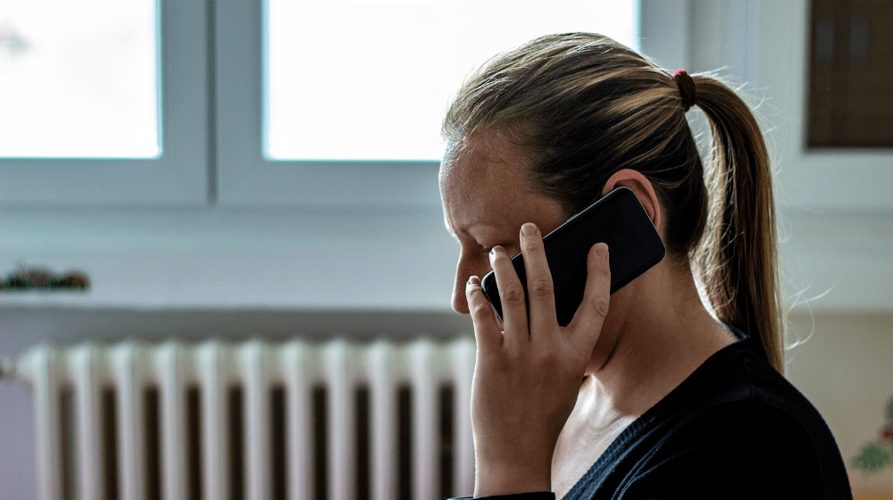 Close up photo of a woman with blonde hair tied in a ponytail sitting in a room and holding a phone against her ear, covering most of her face.