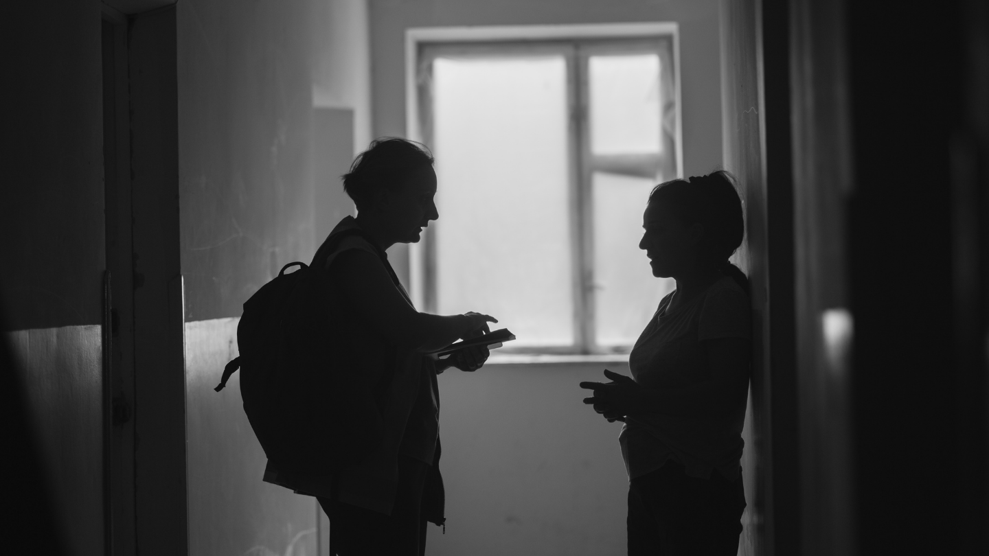 Black-and-white photo of two people standing in a hallway facing each other speaking.