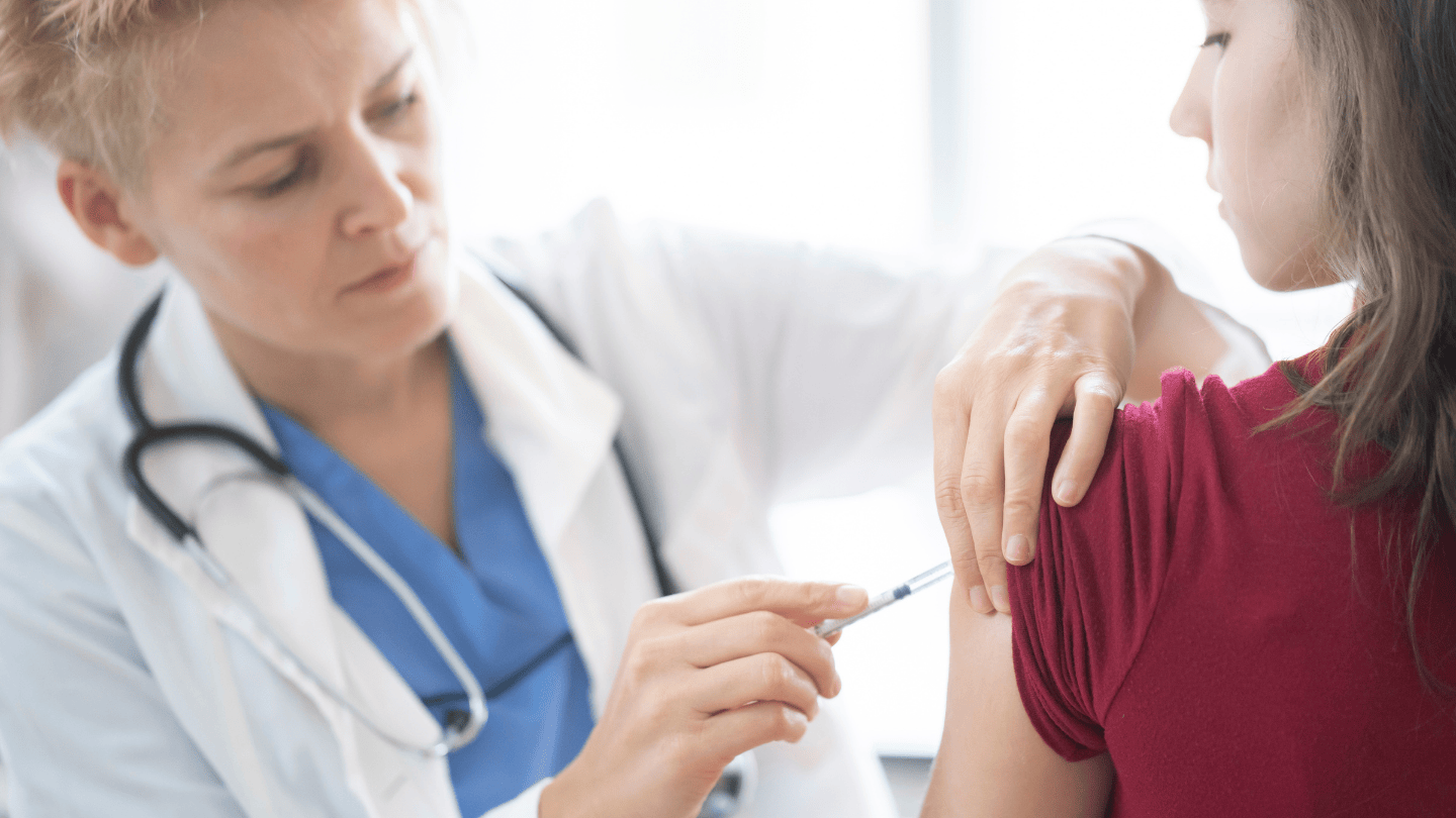 Female health worker wearing white lab coat, blue scrubs and stethoscope around her neck administers vaccine shot to a young woman in a red shirt whose back is turned to the camera but face is turned to the left looking at her exposed arm as the needle is injected