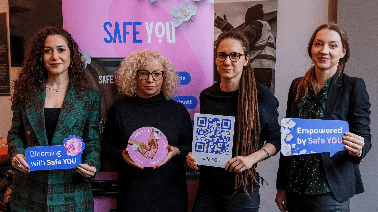 Four women stand together in front of a SafeYOU banner, each one holding a small sign advertising the SafeYOU app