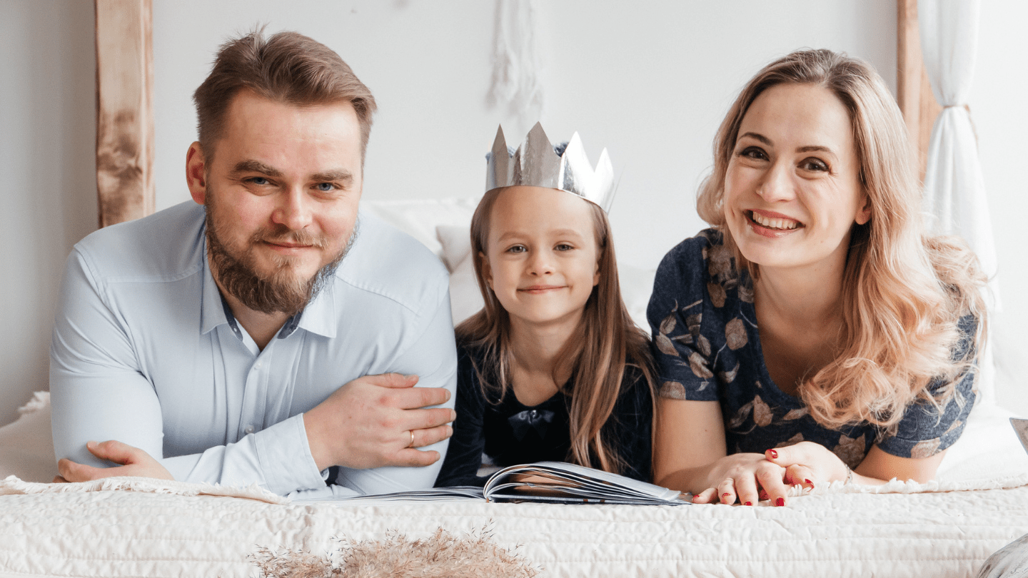 A man, woman, and young girl laying on a bed together reading a book. They are smiling into the camera and the girl has a silver crown on. 