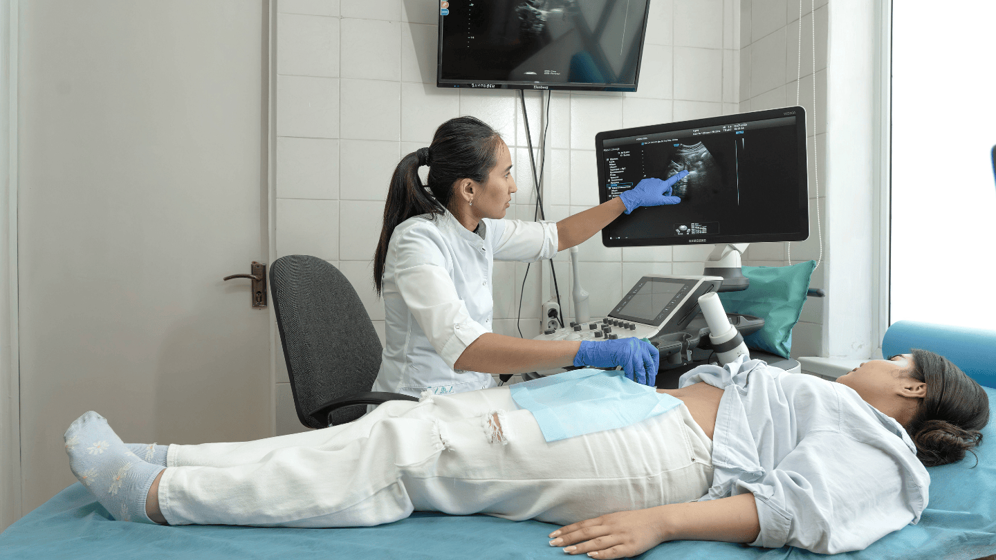 Inside a doctor's exam room, a pregnant young woman in white jeans and a light linen blouse lies on a bed with her face turned away from the camera. Her shirt is lifted to expose her stomach covered in gel. Beside her, a female health worker sits in a chair holding an ultrasound scanner to the pregnant woman's stomach and pointing upward at a monitor showing the ultrasound image. 