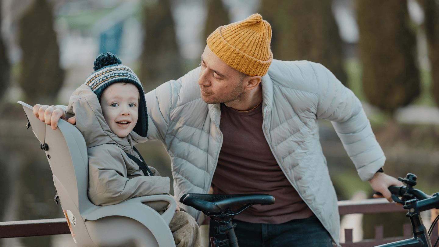 Father and young son outside in a park. Father is wearing a yellow knit hat and light grey quilted jacket. He is holding a bicycle and leaning toward his son, who is sitting in child seat attached to the back of the bicycle. The toddler son is smiling toward the camera, wearing a knit navy hat with a yellow and white pattern and a hooded winter jacket. 