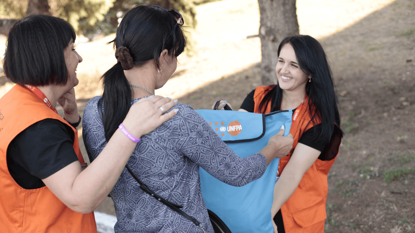 A young woman with dark hair pulled in a ponytail has her back to the camera. On either side of her are two women in bright orange UNFPA vests. The woman in front is handing over a light blue UNFPA Dignity Kit bag. A woman behind lays a comforting hand on the young woman's shoulder.