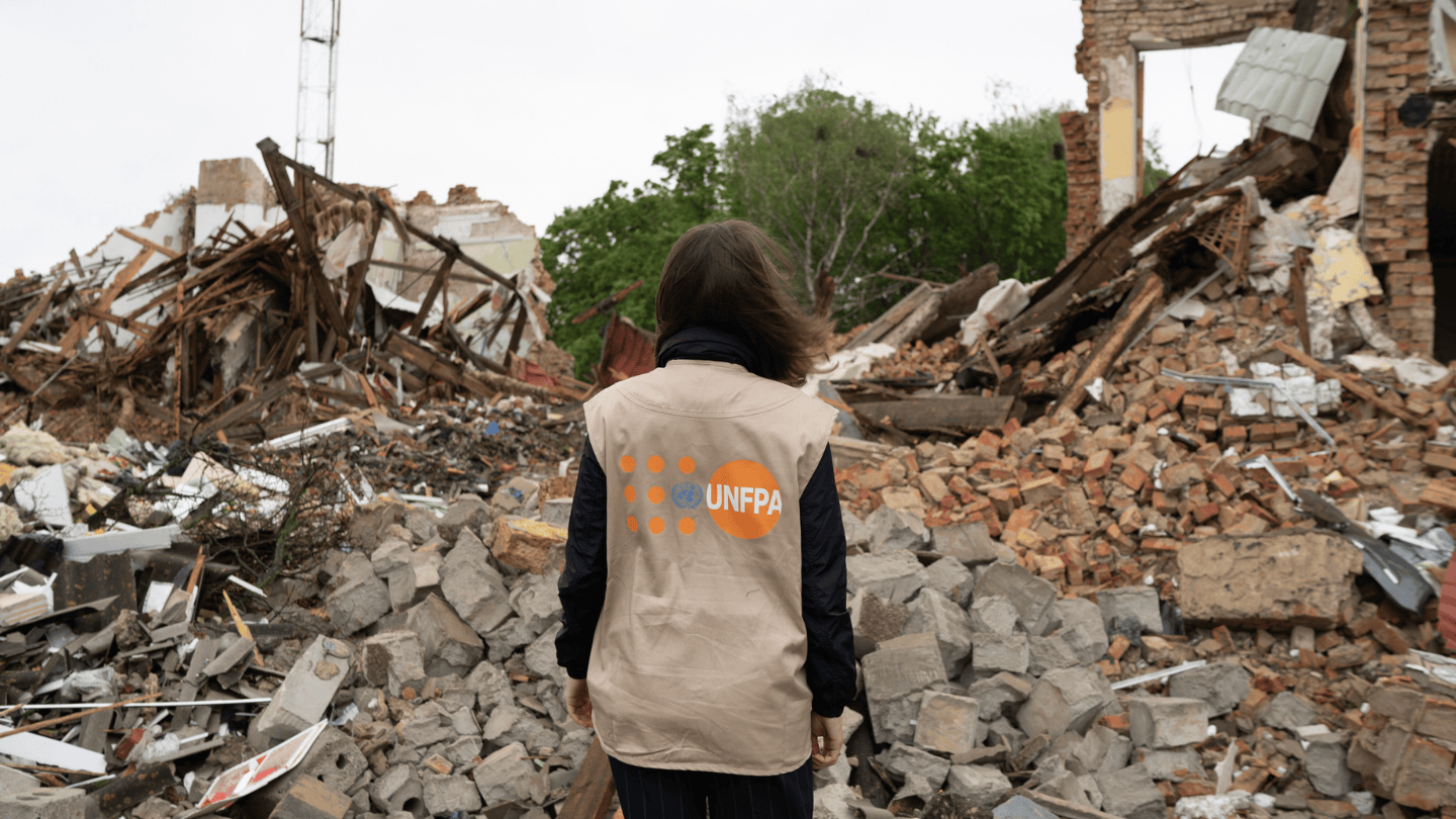 Woman wearing a beige UNFPA vest stands with her back to the camera. In front of her is the rubble of a building completely destroyed by an attack.