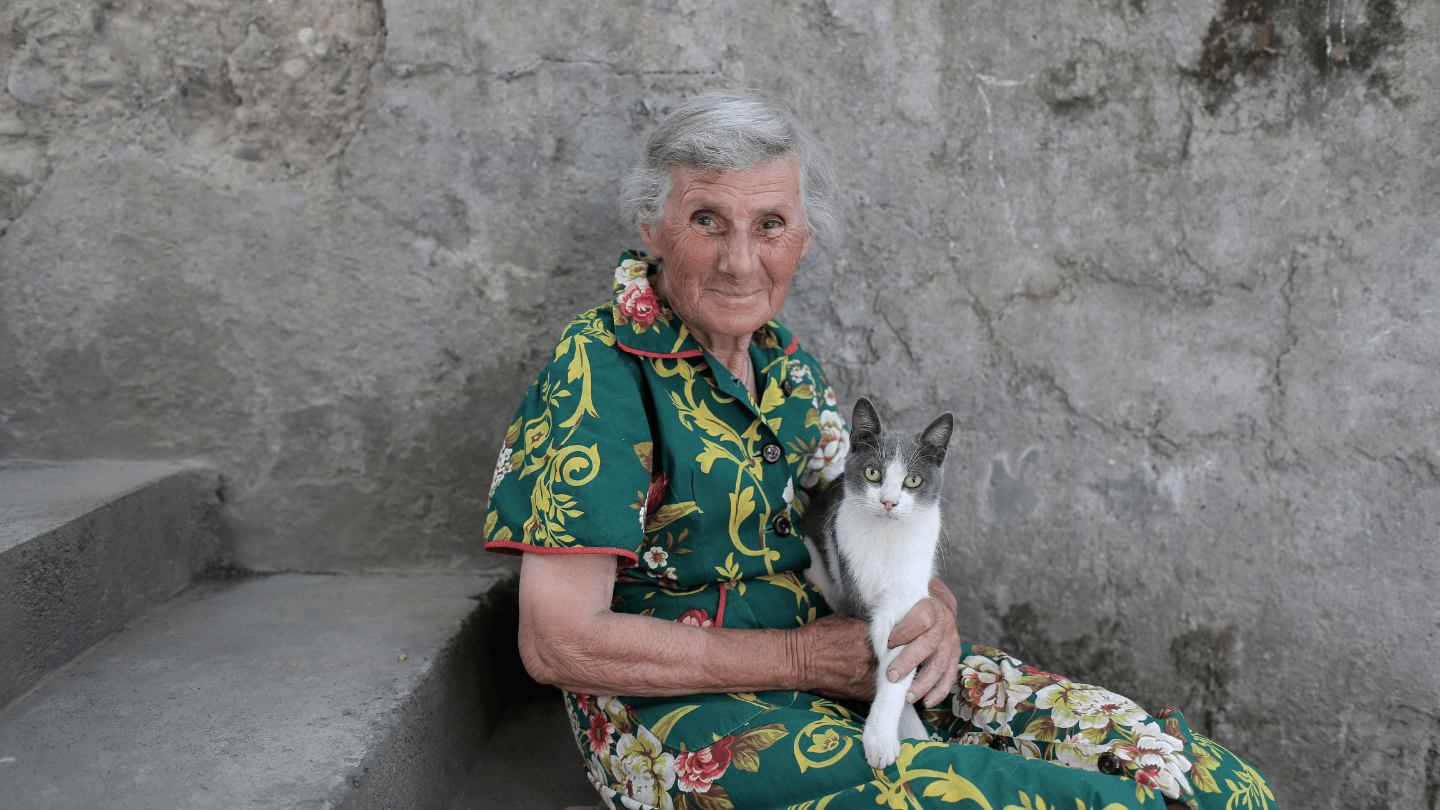 An older woman in a green dress is sitting on stairs with a cat on her lap. She is looking into the camera and smiling.