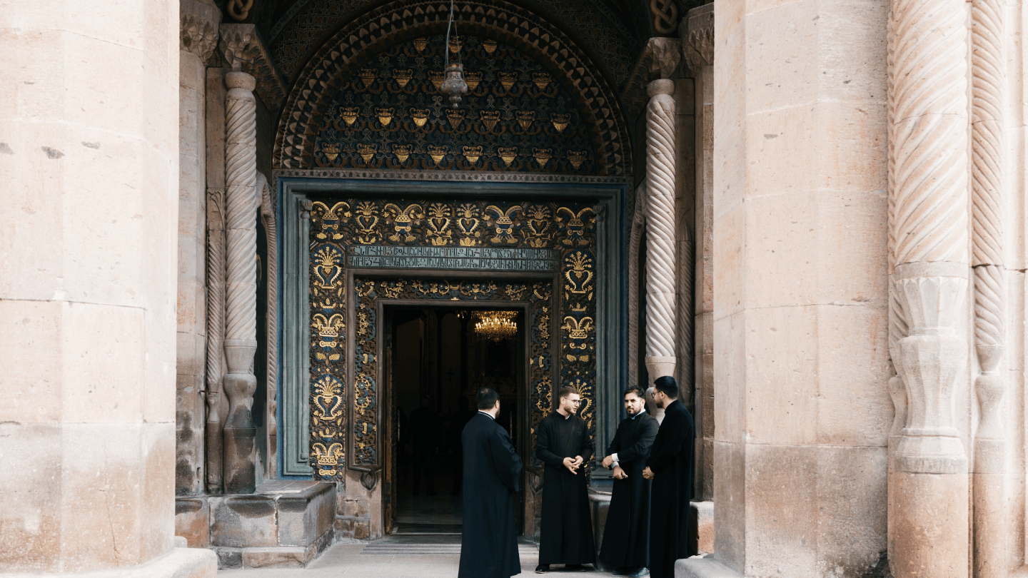 Four men in black religious robes stand in a circle in front of a large ornate church door