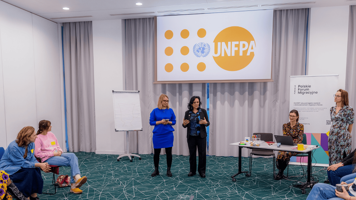 Two women stand in front of a conference room speaking to the audience. Women seated around them listen attentively. A large UNFPA logo is projected on the screen behind them. 