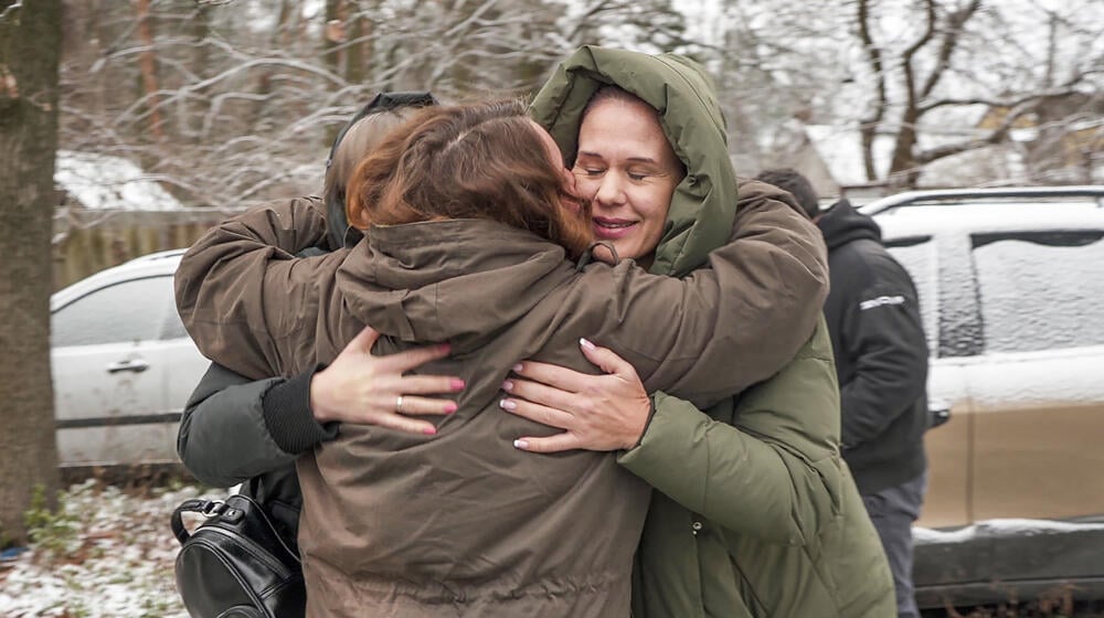 A group of 3 women hug. They are outdoors and there is snow in the background.